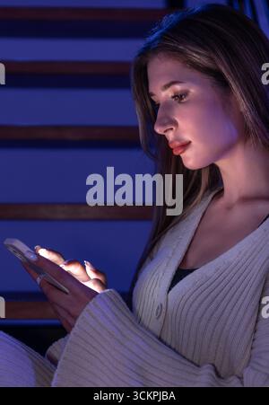 Junge Frau, die auf dem Smartphone auf der Treppe ein Profil schreibt. Blaues Neon. Stockfoto