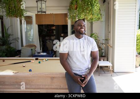 afroamerikanischer Mann, der sich auf Billardtisch auf der Veranda stützt, umgeben von Topfpflanzen und Pool Stockfoto