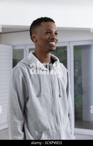 afroamerikanischer Mann in grauem Hoodie stehend und lächelnd im Wohnbereich durch Glasschiebetür Stockfoto