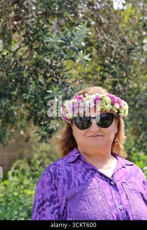 Frau mittleren Alters in lila Bluse mit Sonnenbrille und Blumenkrone draußen im Frühling, natürlichem grünem Hintergrund, saisonales Porträt. Stockfoto