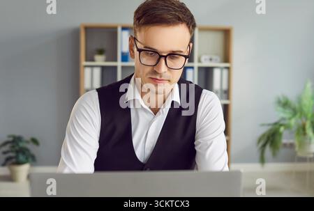 Ernsthafter junger Geschäftsmann mit Brille, der im Büro an einem Laptop arbeitet Stockfoto