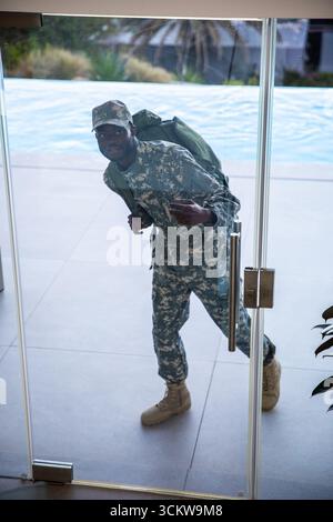 afroamerikanischer Soldat steht an der Glastür in Tarnuniform mit Rucksack auf dem Pooldeck Stockfoto