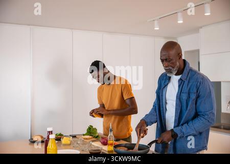 afroamerikaner Senior Vater, der Pfanne rührt, Sohn schneidet Gemüse an Bord auf der Kücheninsel Stockfoto