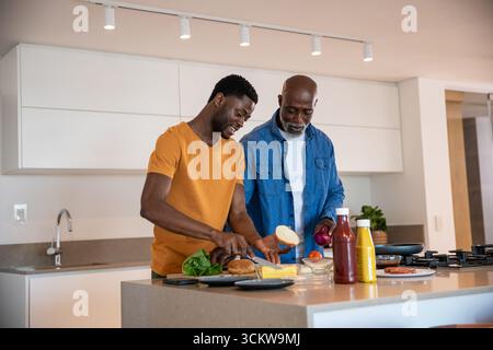 afroamerikanischer Vater und Sohn schneiden Brötchen, um Käsesalat auf der Insel zu arrangieren, in der Pfanne, in der Küche Stockfoto