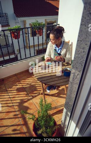 Eine Frau auf einem kleinen Balkon wickelt ein Geschenk an einen Holztisch. Sonnenlicht wirft komplizierte Schatten, und Pflanzen sorgen für grünes Grün. Die Umgebung vermittelt Wärme, Rela Stockfoto