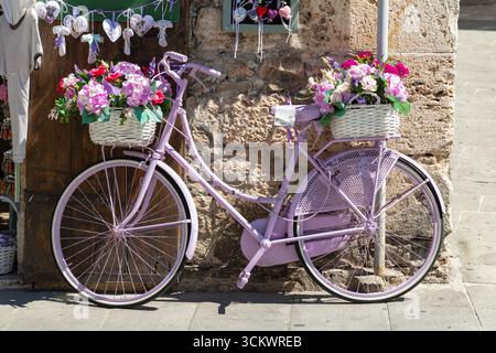 Das charmante lila Fahrrad mit Blumenkörben liegt an einer Steinmauer und verleiht den malerischen Straßen von Assisi, Italien, einen Hauch von Farbe Stockfoto