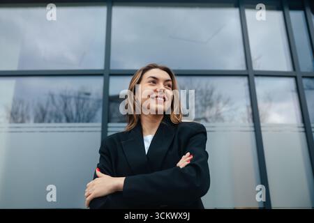 Eine selbstbewusste junge Frau, die mit überkreuzten Armen lächelt, steht vor einem modernen Gebäude und hebt die zeitgenössische Architektur und den urbanen Lebensstil hervor Stockfoto