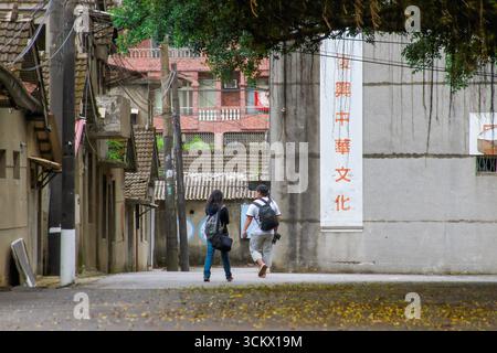 Taoyuan City, Taiwan - 01. Juni 2012: Zwei Menschen mit Rucksäcken gehen auf einer schmalen Straße im Militärdorf Xian Guang entlang. Ein großes Schild mit Stockfoto