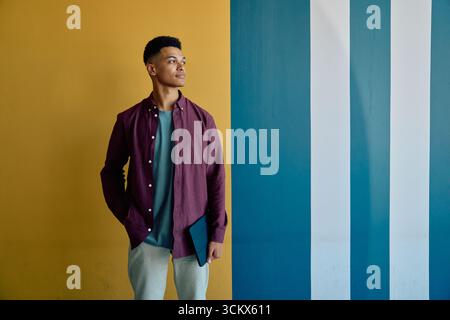Nachdenklicher junger Geschäftsmann in eleganter Freizeit mit Tablet-PC, das im Büro an der Wand steht Stockfoto