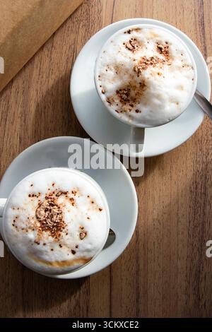 Blick von oben auf zwei Tassen Cappuccino mit Milchschaum und Kakaopulver, serviert in weißen Tassen auf Holztisch. Konzept von Morgenkaffee, gemütlichem Café und Leben Stockfoto