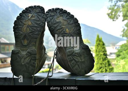 Nahaufnahme der schmutzigen Fußsohlen von Wanderschuhen, die nach einer anstrengenden Wanderung auf einem Balkongeländer ruhen, mit Blick auf eine Berglandschaft. Stockfoto