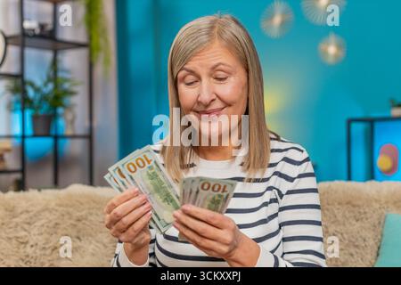 Glückliche kaukasische reife Frau zählt Geld-Dollar-Bargeld in Händen, nachdem sie einen Lottobonus gewonnen oder ein Geschenk erhalten hat. Großmutter lächelt breit auf dem Sitzen Stockfoto