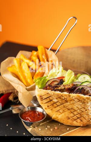Gegrilltes Pita-Sandwich mit Pommes frites und Ketchup auf Holzbrett, leuchtend orange Hintergrund Stockfoto