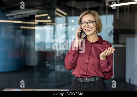 Glückliche Geschäftsfrau mit einer stilvollen Brille, die sich in einem Gespräch mit dem Mobiltelefon einlässt, selbstbewusst lächelt und in einem modernen Büro mit urbanem Hintergrund steht Stockfoto