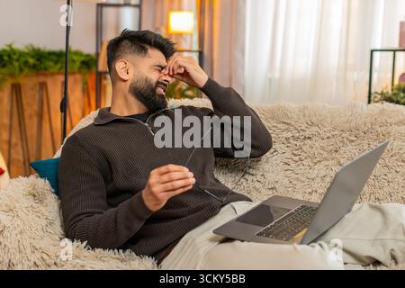 Ein indischer Mann zu Hause schrubbt die Augen mit müdem Ausdruck, während er ein Notebook benutzt, das durch eine lange Bildschirmbelichtung ermüdet ist. Ein Araber auf dem Sofa zeigt Unwohlsein und Augenanstrengung und kämpft mit digitalem Überlastungsstress Stockfoto