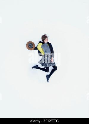 Schöne junge Frau, die auf weißem Schneeteppich neben dem Hut liegt. Oben erhobene Drohnenansicht des Modemodells, das im weißen Schnee bezahlt und entspannt. Stockfoto