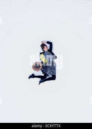 Schöne junge Frau, die auf weißem Schneeteppich neben dem Hut liegt. Oben erhobene Drohnenansicht des Modemodells, das im weißen Schnee bezahlt und entspannt. Stockfoto