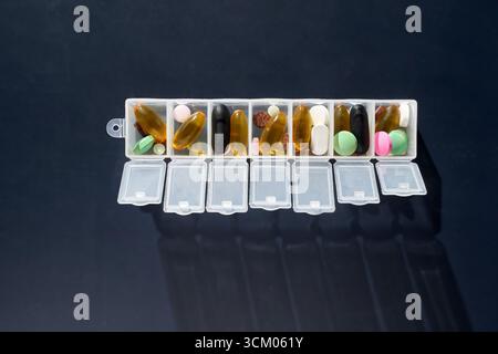 Wöchentlicher Pillen-Organisator mit verschiedenen Kapseln und Tabletten auf dunklem Hintergrund zur Medikamentenbehandlung. Stockfoto