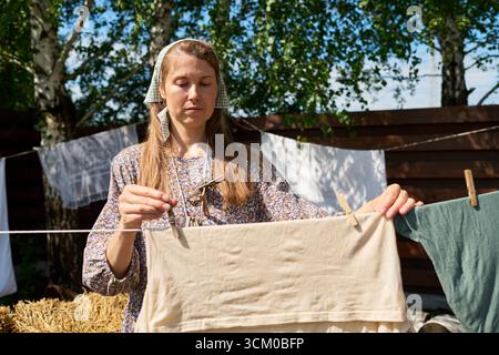 Kaukasische junge Erwachsene Frau, die im Freien Wäsche an der Wäscheleine hängt, im Sonnenlicht mit Bäumen und Holzzaun im Hintergrund steht, Kopftuch und gemustertes Kleid trägt Stockfoto
