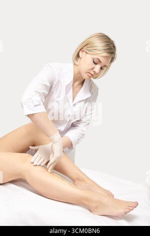 Kaukasische Kosmetikerin, die Wachs auf die Beine junger Frauen zur Haarentfernungsbehandlung im Spa-Setting aufträgt. Stockfoto
