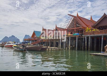 PHUKET, THAILAND - 12. NOVEMBER 2017: Traditionelle thailändische Pfahlhäuser über kristallblauem Wasser mit Booten und bergigem Horizont unter bewölktem Himmel. Stockfoto