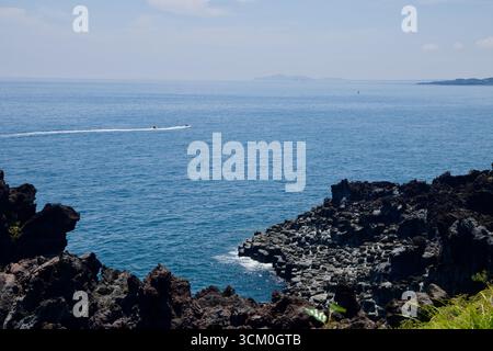 Ein Schnellboot fährt vor der Küste vorbei, während Basaltsäulen der Daepo Jusangjeolli Cliffs über die Wellen ragen. Stockfoto