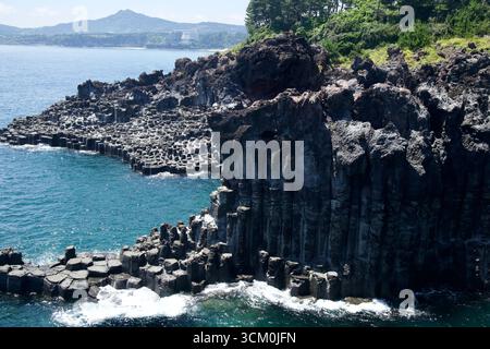 Die Jusangjeolli Cliffs erstrecken sich bis ins Meer und ein entferntes Resort Hotel ist vor der Bergkulisse von Jeju zu sehen. Stockfoto