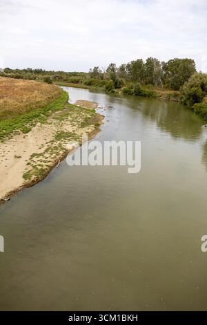 Friedlicher Blick auf einen ruhigen Fluss, der sich durch grasbewachsene Felder und Bäume unter bewölktem Himmel schlängelt Stockfoto