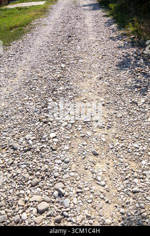 Raue Schotterstraße, die sich durch eine ländliche Landschaft unter Sonnenlicht erstreckt. Stockfoto