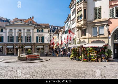 Ein Blick auf den Munzplatz mit Augustinerbrunnen, einem schönen Blumenladen und Schweizer Fahnen im Hintergrund, in der Züricher Altstadt, Schweiz Stockfoto