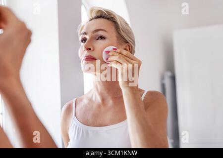 Reife Frau 50 Jahre Hautpflege mit Wattepad, stehend am Spiegel zu Hause. Elegante Seniorin, die tägliche Schönheitsroutine macht, natürliches Altern A Stockfoto