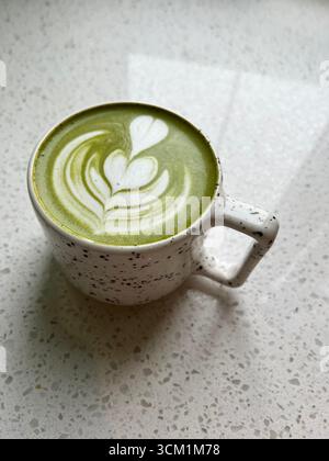 Cremiger grüner Matcha-Tee mit wunderschöner Latte Art in handgemachter Tasse. Aufgenommen auf minimalistischem, gesprenkelten Hintergrund mit weichem Tageslicht, perfekt für Kaffees Stockfoto