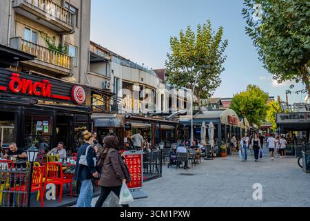 Die Leute sitzen in Cafés im Freien und laufen entlang einer belebten Straße, die von Restaurants und Geschäften gesäumt ist. Adapazarı, Sakarya, Türkiye – 09. September 2025. Stockfoto