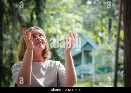 Junge Frau blickt mit einem fröhlichen Ausdruck auf und erhob die Hände in einem üppigen Wald in der Nähe einer gemütlichen Holzhütte Stockfoto