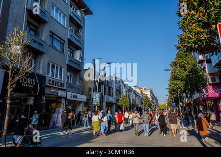 An einem sonnigen Tag laufen Menschenmassen entlang einer breiten Fußgängerzone, die von Geschäften und Geschäften gesäumt ist. Adapazarı, Sakarya, Türkiye – September Stockfoto