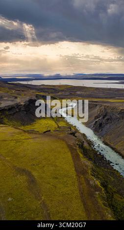 Aus der Vogelperspektive eines sich windenden Flusses durch einen moosbedeckten Canyon mit vulkanischen Felsen, einem fernen See und sonnendurchfluteten Wolken in Island. Stockfoto