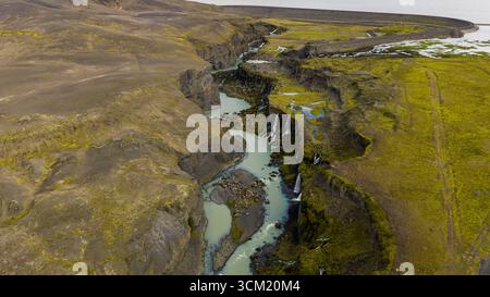 Ein sich windender türkisfarbener Fluss fließt durch einen zerklüfteten Canyon mit Wasserfällen, die von steilen Klippen umgeben sind, umgeben von vulkanischem Felsen und grünem Moos. Stockfoto