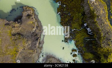 Türkisfarbener Fluss fließt durch zerklüftetes vulkanisches Gelände in Island, flankiert von moosbedeckten Klippen und kleinen kaskadierenden Wasserfällen in einer perspektivischen Perspektive Stockfoto