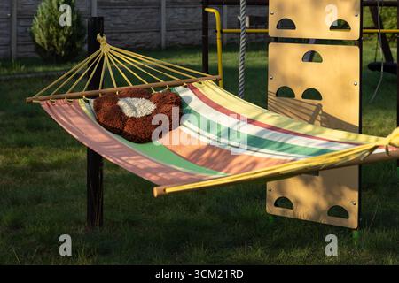Bunt gestreifte Hängematte mit flauschigem Kissen im gemütlichen Hinterhof des Landhauses. Entspannung im Freien, Garten Freizeit und ländliches Leben Stockfoto