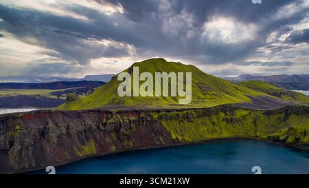 Aus der Vogelperspektive auf einen mit grünem Moos bedeckten vulkanischen Berg in Island mit einem tiefen blauen Kratersee, umgeben von rauem Gelände und einem wolkengefüllten Himmel. Stockfoto