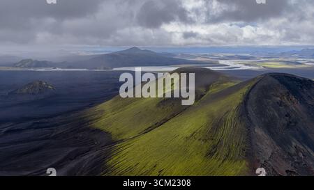 Aus der Vogelperspektive auf Islands vulkanische Landschaft mit moosbedeckten Hügeln, einem geflochtenen Flusssystem und einem bewölkten Himmel, der eine stimmungsvolle Atmosphäre schafft. Stockfoto