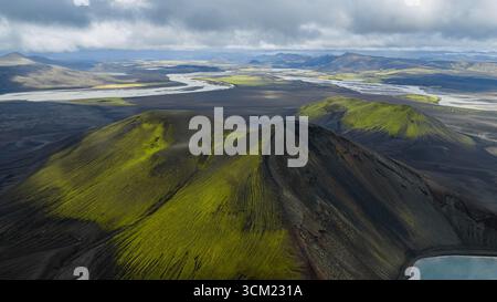 Aus der Vogelperspektive sehen moosbedeckte vulkanische Hügel, einen sich windenden Fluss und einen kleinen See unter einem bewölkten Himmel in Islands rauem Gelände. Stockfoto