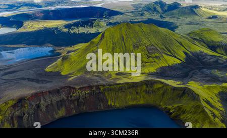 Aus der Vogelperspektive auf einen moosbedeckten vulkanischen Krater mit einem lebhaften blauen See an der Basis, umgeben von dunklem Gelände, Flüssen und zerklüfteten Hügeln in Island. Stockfoto