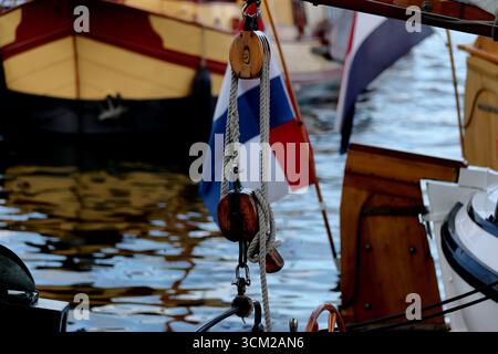 Takelage mit gepaarten Holzblöcken auf einem Segelschiff bei Sail 2025 in Amsterdam, Niederlande, mit niederländischer Flagge und kleinen Booten auf reflektierendem Wasser. Stockfoto