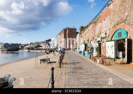 Malerischer Blick auf den Fluss exe auf den Quay in exeter devon mit historischen Steinhäusern, Geschäften und Cafés entlang einer gepflasterten Uferpromenade unter blauem Himmel Stockfoto