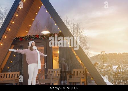 Eine Frau freut sich über die Sonne und einen neuen Tag an der Schwelle ihres hölzernen Hauses in der Natur und breitet ihre Arme weit aus. Neujahr und ein neuer Tag im Winter Stockfoto