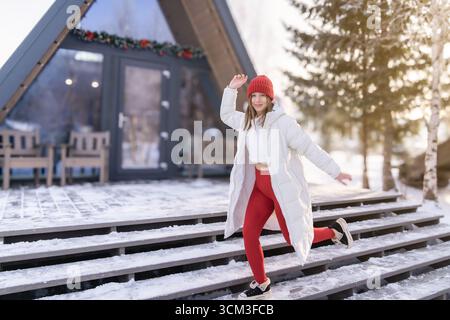 Junge Frau, die den Wintertag vor einer gemütlichen Hütte im Schnee genießt, während sie einen weißen Mantel und einen roten Hut trägt. Stockfoto
