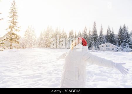 Fröhliche junge Frau, die an einem sonnigen Wintertag in einer verschneiten Waldlandschaft mit Bäumen im Hintergrund den Schneefall genießt. Stockfoto