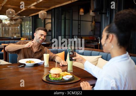 Ein vielseitiges Paar mit Feinschmecker genießt ein zwangloses Abendessen und einen Telefonmoment, bei dem sonnige Vibes und Drinks geteilt werden Stockfoto