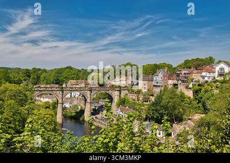Eine malerische Szene zeigt ein Steinviadukt über einem Fluss mit Häusern und üppigen Bäumen am Ufer unter einem hellblauen Himmel in Knaresborough, Großbritannien. Stockfoto
